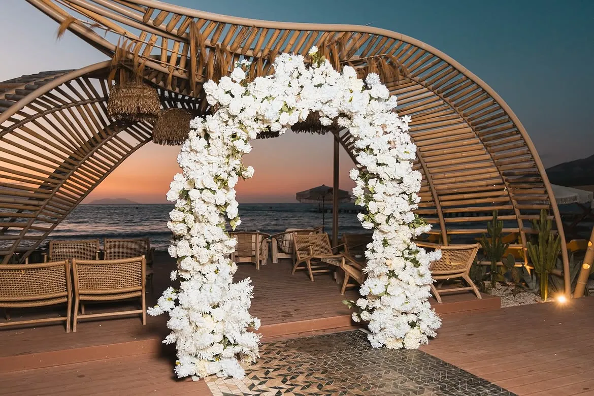 Seaside wedding ceremony with a white floral arch at sunset at BOJO Resort